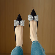 Black high-heeled shoes with large crystal bows worn by a person in blue jeans against a brown background