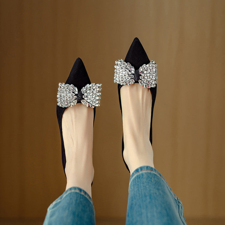 Black high-heeled shoes with large crystal bows worn by a person in blue jeans against a brown background