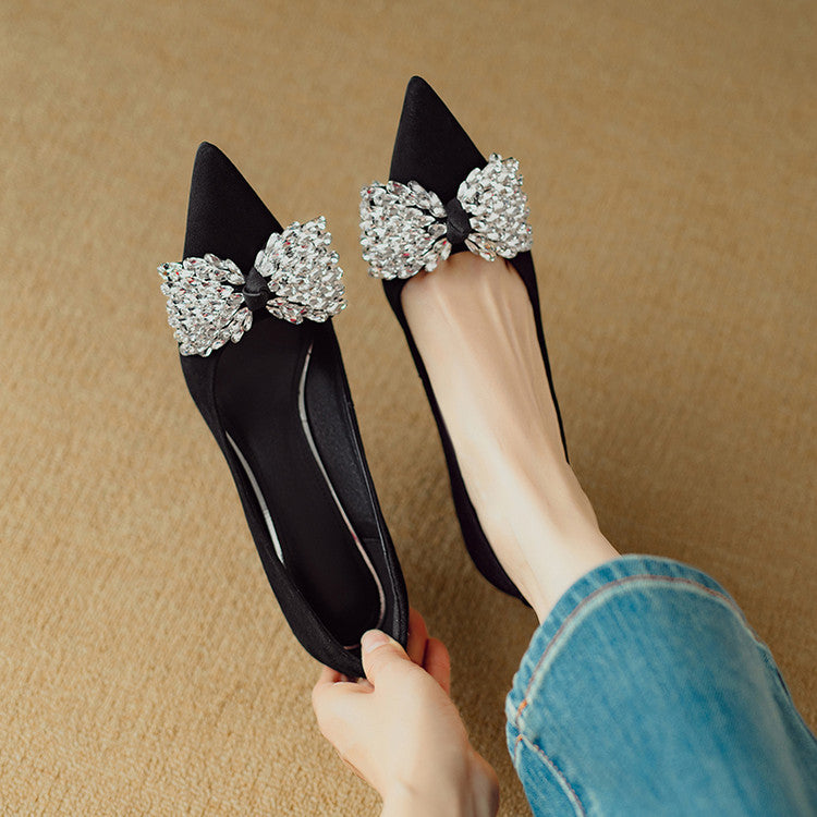 Black flats with large crystal bows held by a person wearing blue jeans.